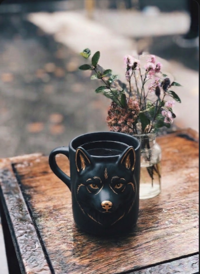 A unique black ceramic mug shaped like a wolf's face, placed on a wooden table with a small vase of flowers in the background.