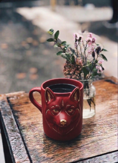 A red wolf-shaped mug filled with coffee sits on a wooden table next to a small vase of dried flowers in a cozy setting.