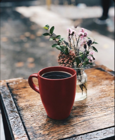 A red coffee mug sits on a wooden table next to a small vase of pink flowers, creating a cozy café atmosphere.
