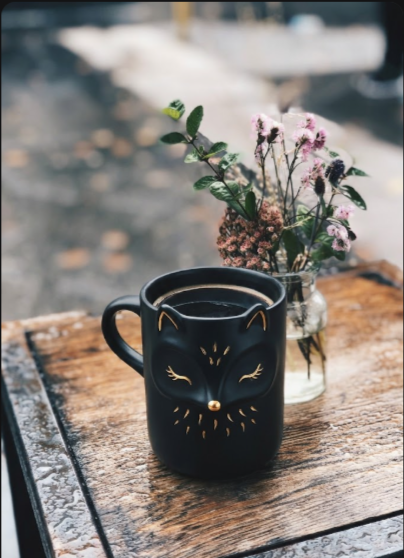 A black cat-shaped mug filled with coffee sits on a wooden table beside a small vase of pink flowers.