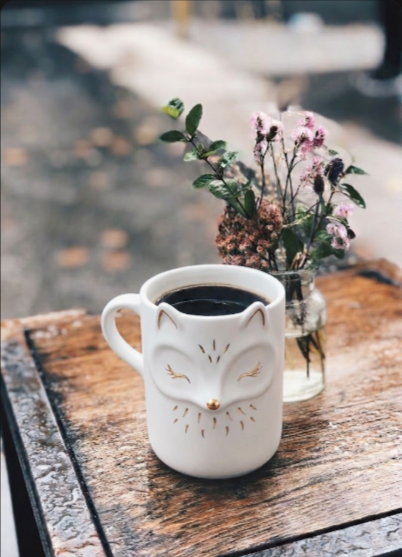 A charming fox-shaped coffee mug filled with black coffee, placed on a wooden table next to a small vase of flowers.