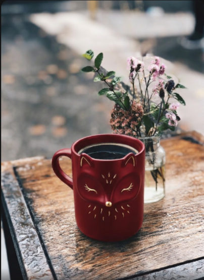 A charming red fox-shaped mug filled with coffee sits on a wooden table beside a small vase of flowers.