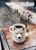 A unique wolf-shaped mug filled with coffee sits on a wooden table beside a small vase of flowers.