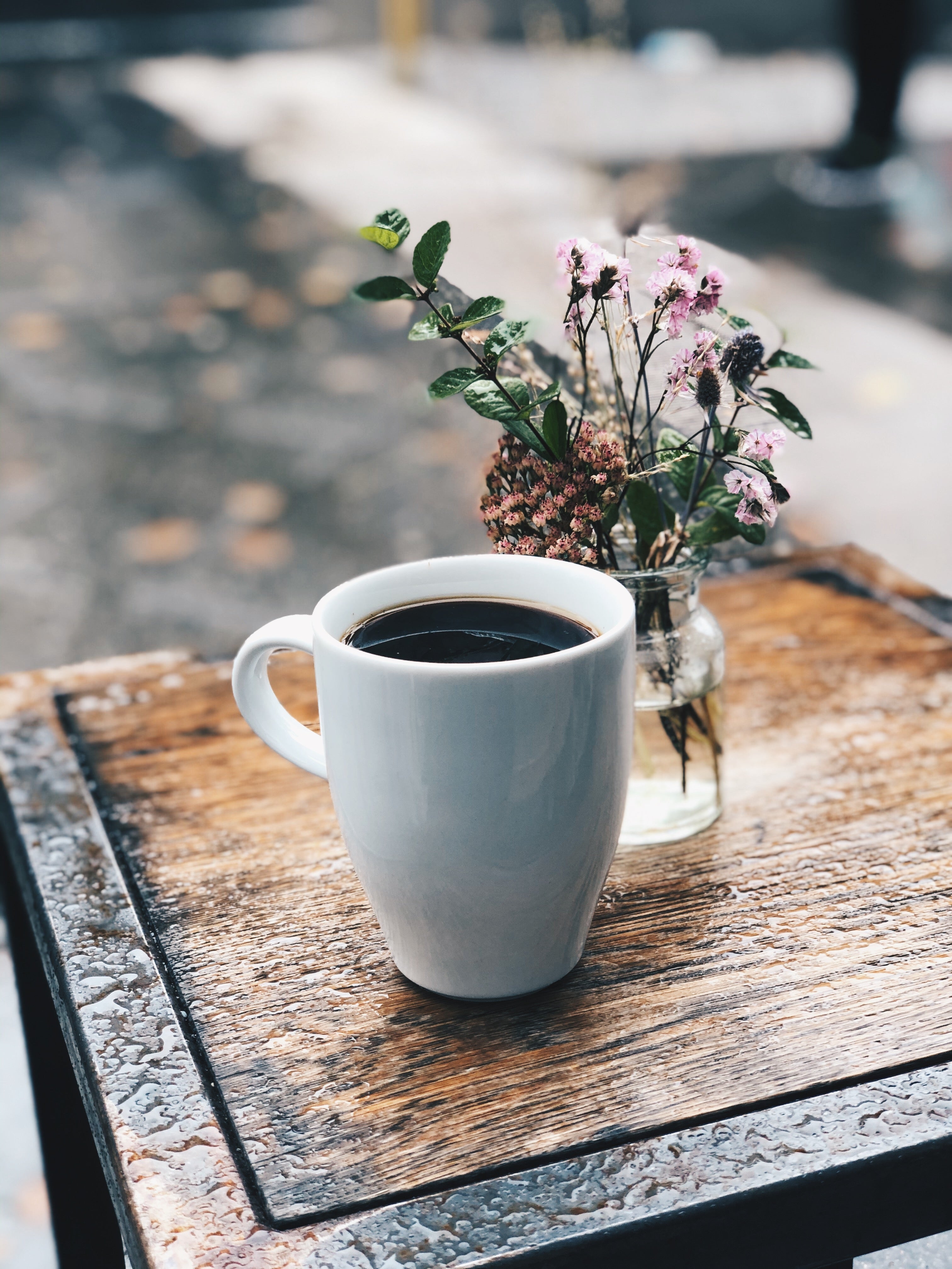 A steaming cup of coffee on a wooden table beside a small vase with fresh flowers, creating a cozy atmosphere.